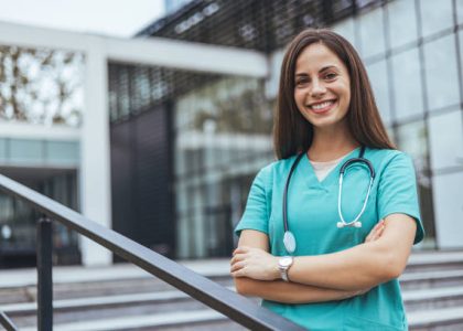 istockphoto-2155050537-612×612 A smiling Caucasian healthcare worker stands confidently outdoors with arms crossed, wearing medical scrubs and a stethoscope, symbolizing professional medical care.