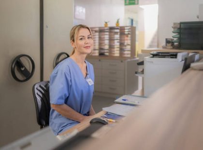 istockphoto-2198864708-612×612 Caucasian female veterinarian university clinic receptionist in blue scrubs, working professionally in a busy medical environment, maintaining patient records and assisting visitors effectively.